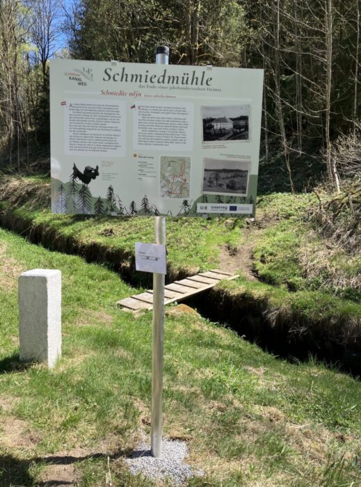 Infotafel „Schmiedmühle“ an einem Waldweg neben einem schmalen Bach mit kleiner Holzbrücke; Text, Karte und Bilder erklären die Geschichte des Ortes.