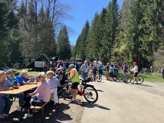 Viele Menschen und Fahrräder auf breitem Waldweg; Sitzbänke und Tische laden zur Rast ein. Informelle Veranstaltung oder Treffpunkt an sonnigem Tag im Grünen.
